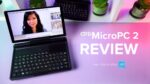 A dynamic thumbnail for a GPD MicroPC 2 review video. A black GPD MicroPC 2 is prominently featured in the foreground, open and angled towards the viewer, with its screen displaying a video conference call featuring a woman's face. The laptop's compact keyboard and trackpad are visible. The background is a white shelf with potted plants and another GPD MicroPC 2. The desk below the laptop is a vibrant pink and purple gradient. Large white text to the right of the laptop reads "GPD MicroPC 2 REVIEW", and smaller text below that says "Intel N250 vs N300" alongside the "Intel inside" logo.