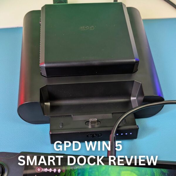 A close-up photograph of the black GPD WIN 5 Smart Dock sitting on a light blue surface. The dock is designed to hold the GPD WIN 5, with the console's screen partially visible in the foreground, displaying a gaming interface. The dock itself is connected by a black cable, and its LED indicator lights are visible near the charging port. Large white text overlaid on the image reads, "GPD WIN 5 SMART DOCK REVIEW."