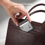 Tuvplānā redzama roka, kas ievieto sudraba daudzportu lādētāju brūnas ādas rokassomas sānu kabatā, fonā redzams teksts "Multi-port Charging, Worry-free Travel".